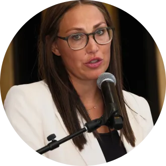 A woman with long brown hair and glasses speaks into a microphone. She is wearing a white blazer over a dark top.