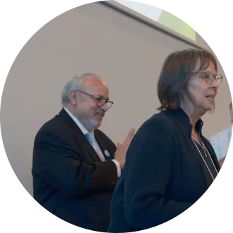 Man in black suit with light-blue tie stands behind woman in darker blue collared shirt.