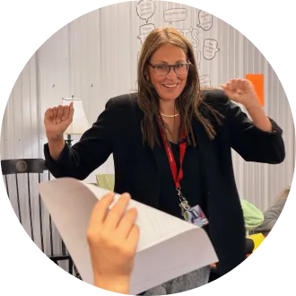 A teacher walks across a classroom, smiling and looks at a student's hand that is raised and holding a sheet of paper.