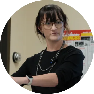 A woman wearing glasses and a black top stands with her arms crossed, a small microphone clipped to her shirt, appearing focused as she listens in a classroom setting with posters on the wall behind her.
