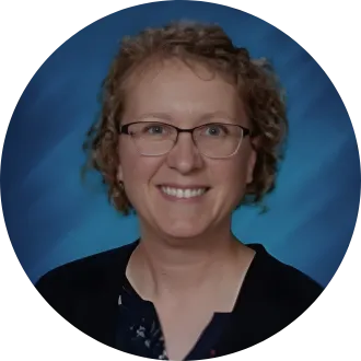 Woman with short, dark blonde hair, glasses, and a black shirt, in front of a blue backdrop.