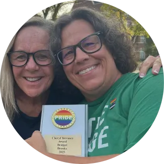 Two smiling women wearing glasses pose closely together outdoors, one in a green T-shirt with a rainbow pin. They hold a plaque with a rainbow “PRIDE” emblem reading “Cheryl Terrance Award — Bridget Brooks — 2025.”