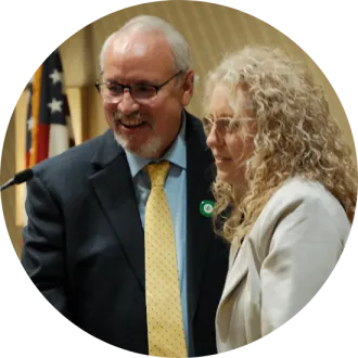 Man in suit with light blue shirt and yellow tie standing next to woman in tan suit jacket, behind a podium.