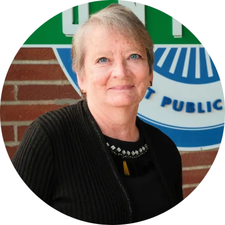 North Dakota United-Retired President Toni Gumeringer standing in front of NDU logo on brick wall in Bismarck. She is shown wearing a black sweater and shirt, smiling at camera.