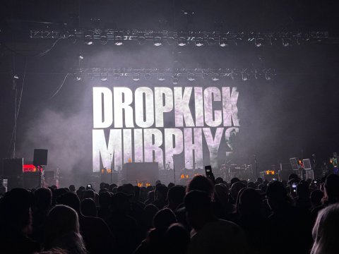 A crowd of people standing in packed concert hall that is dark with the Dropkick Murphys band logo projected on large screen behind empty stage.